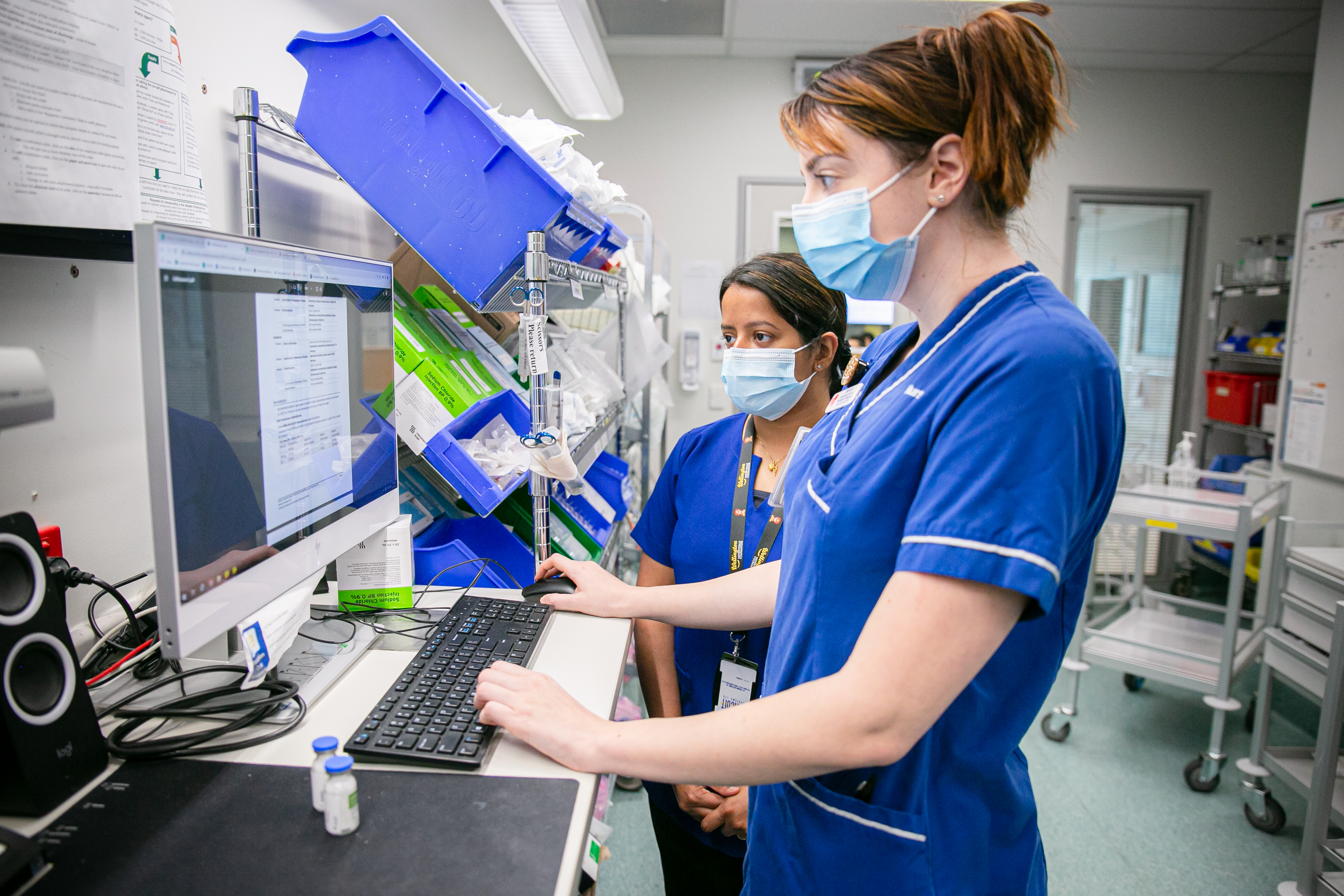Two nurses in clinical uniforms work at a computer workstation in a hospital clinical area, reviewing information on a screen. Shelving with medical supplies and equipment is visible around them, indicating a busy ward or treatment room.