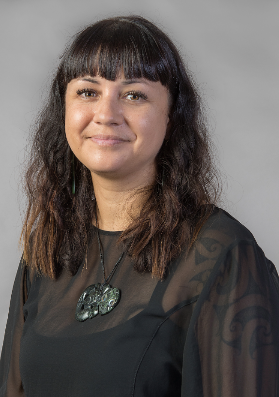 Portrait of Rīpeka Tamanui‑Hurunui wearing a sheer black top and a carved pendant necklace, photographed against a neutral grey background.