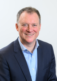 Alex Gordon, head‑and‑shoulders studio portrait, wearing a navy blazer and light‑blue collared shirt against a plain white background.