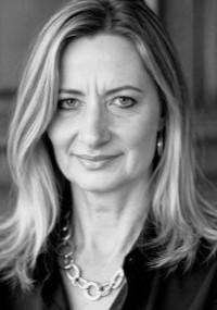 Portrait of Amanda Singleton, photographed in black and white, wearing a dark jacket and a chain necklace, with shoulder‑length light hair styled neatly against a studio backdrop.